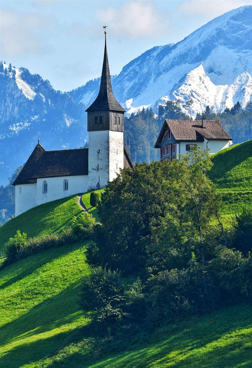 A white chapel with a dark spire on a green hillside, snow-covered alpine peaks rising behind it.